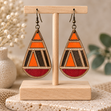 Load image into Gallery viewer, Geometric teardrop hand-painted wooden earrings displayed front-facing on a light wood T-stand, showing orange, deep red, dark accents and bronze-tone hooks.