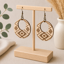 Load image into Gallery viewer, Angled view of Tribal Spirit engraved wooden earrings on a light wood T-stand, highlighting natural wood grain and the fine laser-engraved geometric lines.