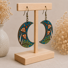 Load image into Gallery viewer, Enchanted Forest Moon wooden earrings shown at an angle on a light wood stand, highlighting the hand-painted design and natural wood construction.

