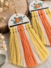 Load image into Gallery viewer, Close-up of Sun Whisper wooden earrings showing the hand-painted floral design, gold-tone hooks, knotted macramé details and textured tassels
