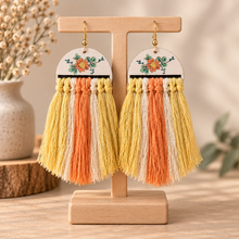 Load image into Gallery viewer, Sun Whisper handmade wooden earrings with hand-painted floral design and cream, yellow and coral macramé tassels on a light wood stand
